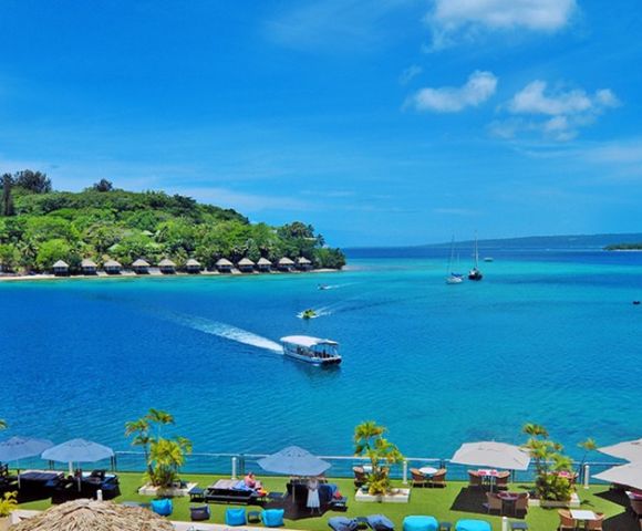 A serene beach scene with vibrant blue water, a boat leaving a wake, distant sailboats, lush greenery, and a clear sky, creating a relaxing tropical atmosphere.