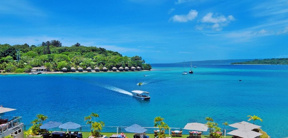 A serene beach scene with vibrant blue water, a boat leaving a wake, distant sailboats, lush greenery, and a clear sky, creating a relaxing tropical atmosphere.