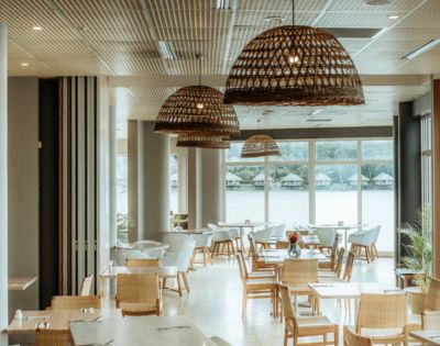 Spacious restaurant with wicker chairs and pendant lights, large windows overlooking a lake. Bright, airy atmosphere with natural decor.