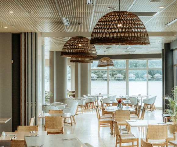 Spacious restaurant with wicker chairs and pendant lights, large windows overlooking a lake. Bright, airy atmosphere with natural decor.