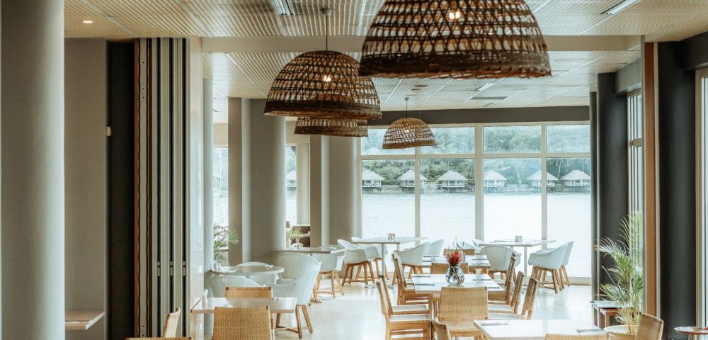 Spacious restaurant with wicker chairs and pendant lights, large windows overlooking a lake. Bright, airy atmosphere with natural decor.