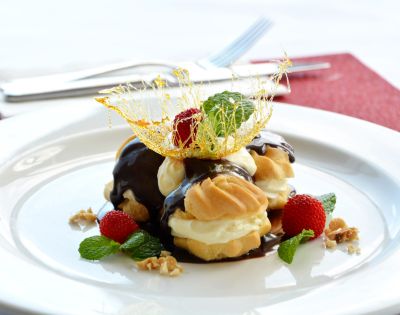 Cream puffs drizzled with chocolate sauce, topped with spun sugar and mint leaves, sit on a white plate surrounded by strawberries and walnuts.