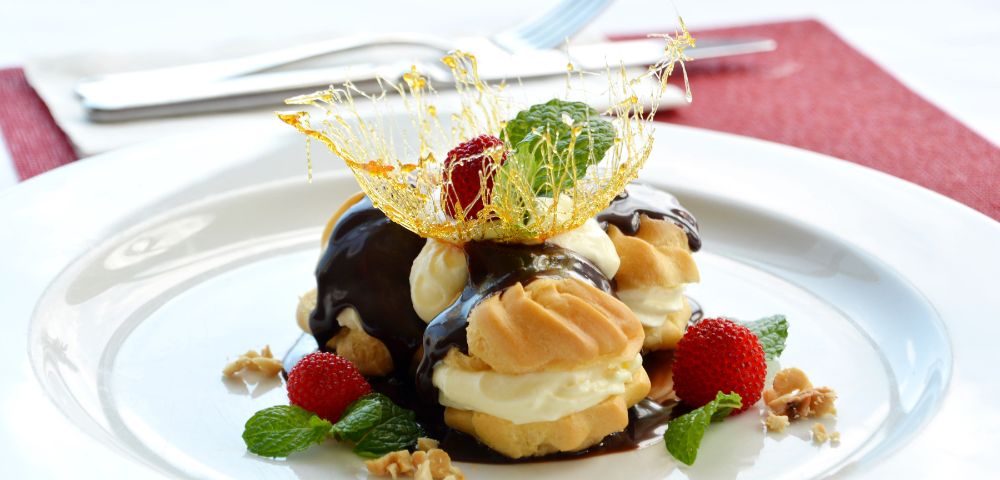 Cream puffs drizzled with chocolate sauce, topped with spun sugar and mint leaves, sit on a white plate surrounded by strawberries and walnuts.