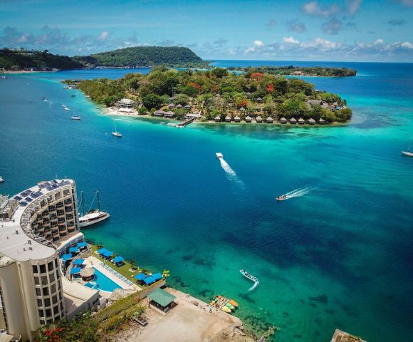 Aerial view of a vibrant turquoise bay with boats leaving white trails, an island with lush greenery, and a coastal hotel complex featuring pools.
