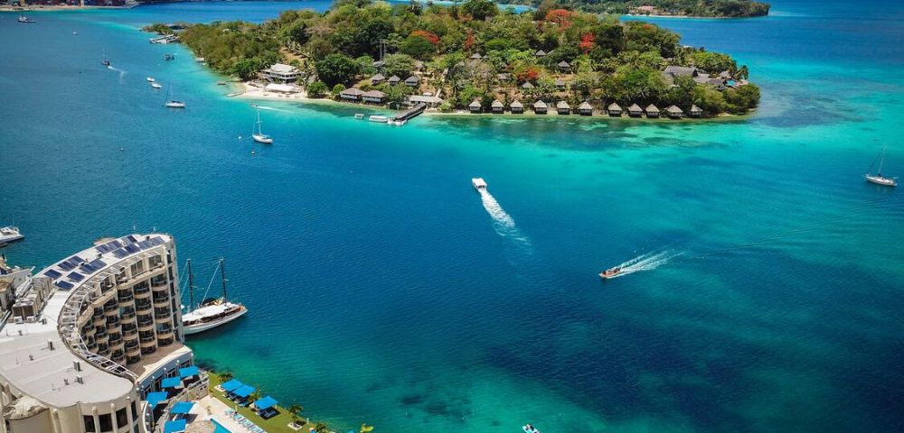 Aerial view of a vibrant turquoise bay with boats leaving white trails, an island with lush greenery, and a coastal hotel complex featuring pools.