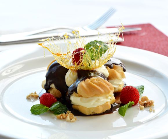 Cream puffs drizzled with chocolate sauce, topped with spun sugar and mint leaves, sit on a white plate surrounded by strawberries and walnuts.