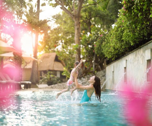 A woman in a blue swimsuit lifts a child joyfully in a serene pool surrounded by lush greenery. Pink flowers blur the foreground, enhancing the warm ambiance.