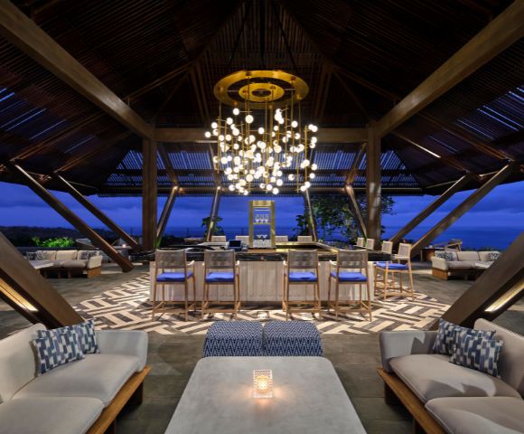 Spacious lounge with modern decor at dusk, featuring cozy seating, a central bar, and an elegant chandelier, creating a warm, inviting atmosphere.