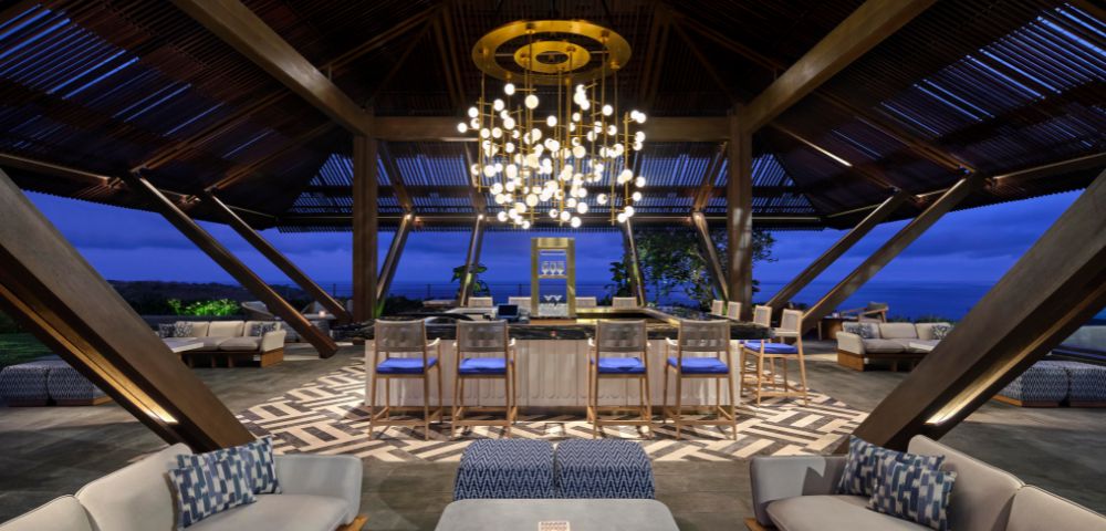 Spacious lounge with modern decor at dusk, featuring cozy seating, a central bar, and an elegant chandelier, creating a warm, inviting atmosphere.