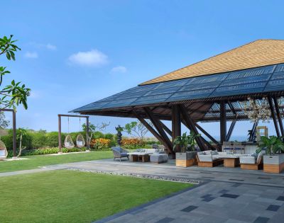 Spacious outdoor lounge with wooden pavilion, surrounded by greenery and hanging chairs. Bright blue sky evokes a serene, tropical atmosphere.