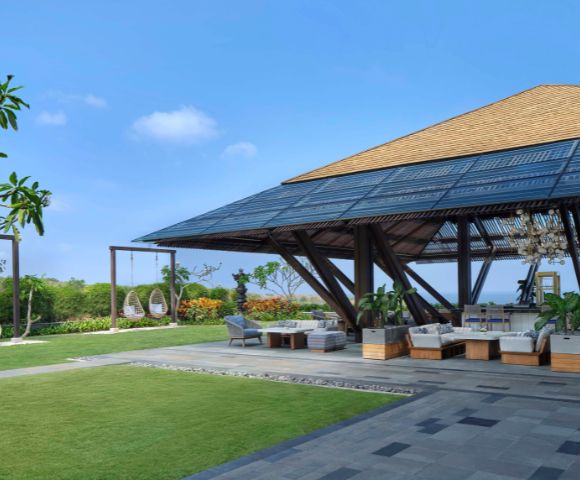 Spacious outdoor lounge with wooden pavilion, surrounded by greenery and hanging chairs. Bright blue sky evokes a serene, tropical atmosphere.
