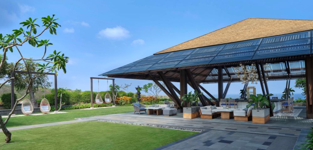 Spacious outdoor lounge with wooden pavilion, surrounded by greenery and hanging chairs. Bright blue sky evokes a serene, tropical atmosphere.