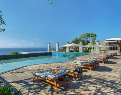 Luxurious poolside with wooden lounge chairs under white umbrellas. Overlooking a serene ocean and lush greenery, exuding a tranquil, sunny ambiance.