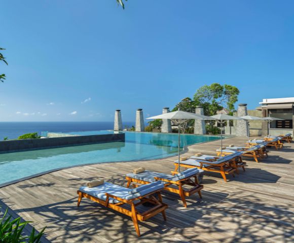 Luxurious poolside with wooden lounge chairs under white umbrellas. Overlooking a serene ocean and lush greenery, exuding a tranquil, sunny ambiance.