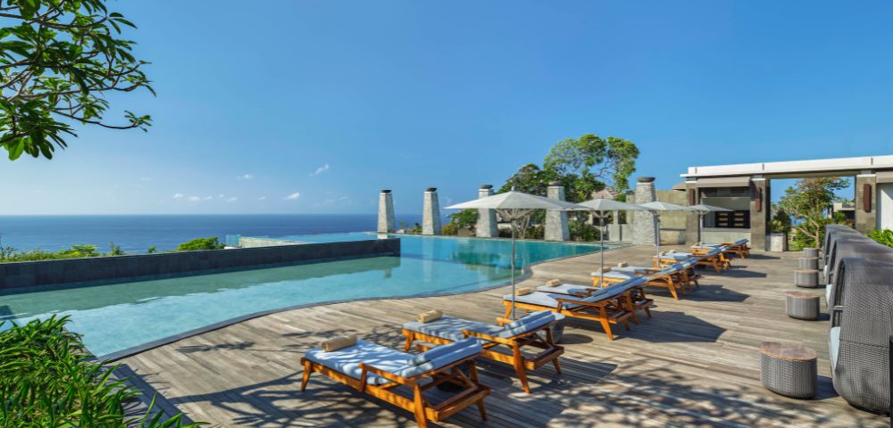 Luxurious poolside with wooden lounge chairs under white umbrellas. Overlooking a serene ocean and lush greenery, exuding a tranquil, sunny ambiance.
