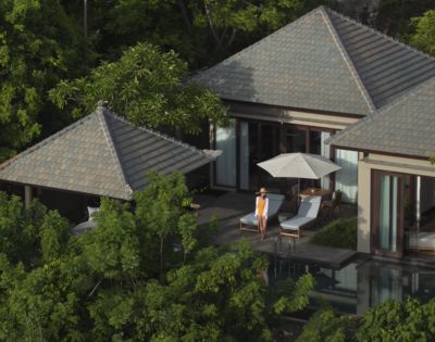 Aerial view of a serene villa nestled among lush trees, featuring a poolside terrace with lounge chairs and an umbrella. A person stands, exuding relaxation.