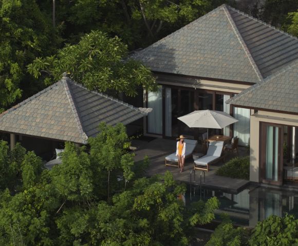 Aerial view of a serene villa nestled among lush trees, featuring a poolside terrace with lounge chairs and an umbrella. A person stands, exuding relaxation.