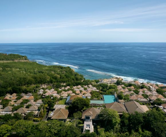 Aerial view of a coastal resort featuring numerous thatched-roof villas surrounded by lush greenery, overlooking a vast, tranquil blue ocean under a clear sky.