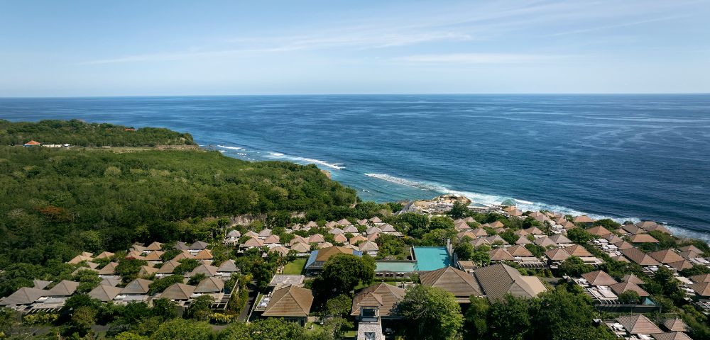 Aerial view of a coastal resort featuring numerous thatched-roof villas surrounded by lush greenery, overlooking a vast, tranquil blue ocean under a clear sky.
