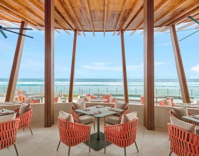 Modern beachfront restaurant with coral-colored wicker chairs and wooden beams. Large windows offer a stunning ocean view under a clear blue sky.