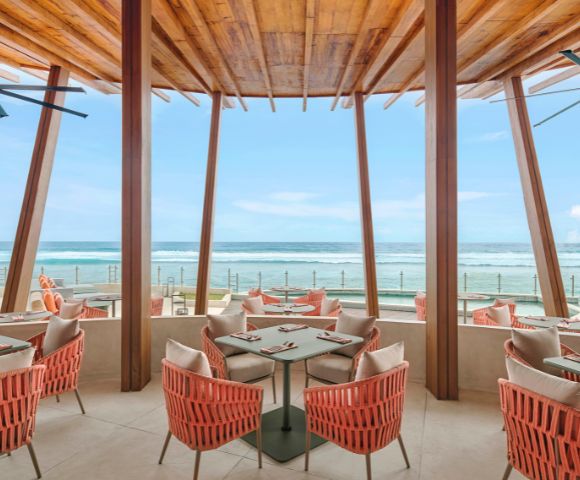 Modern beachfront restaurant with coral-colored wicker chairs and wooden beams. Large windows offer a stunning ocean view under a clear blue sky.