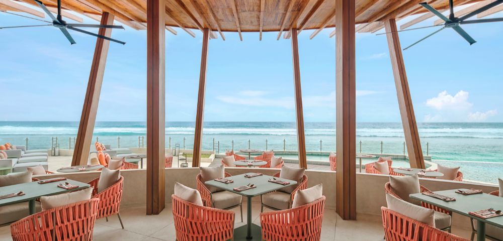 Modern beachfront restaurant with coral-colored wicker chairs and wooden beams. Large windows offer a stunning ocean view under a clear blue sky.