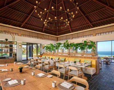 Cozy restaurant with wooden tables, cushioned chairs, and lush greenery. Large windows reveal a bright ocean view. Elegant chandelier overhead.