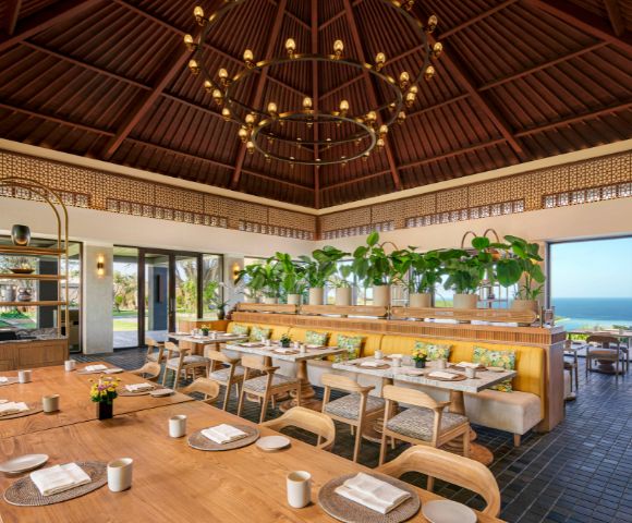 Cozy restaurant with wooden tables, cushioned chairs, and lush greenery. Large windows reveal a bright ocean view. Elegant chandelier overhead.