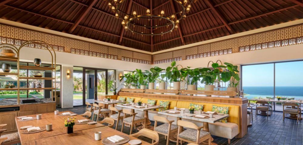 Cozy restaurant with wooden tables, cushioned chairs, and lush greenery. Large windows reveal a bright ocean view. Elegant chandelier overhead.