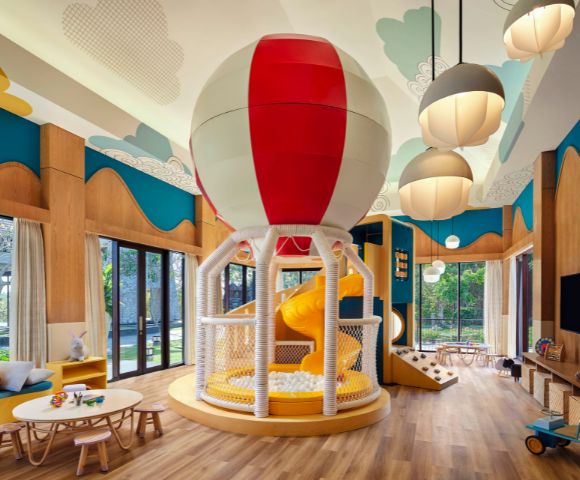 Bright indoor playroom featuring a large balloon-themed slide, colorful decor, wood flooring, round tables, and clouds on the ceiling, creating a playful atmosphere.