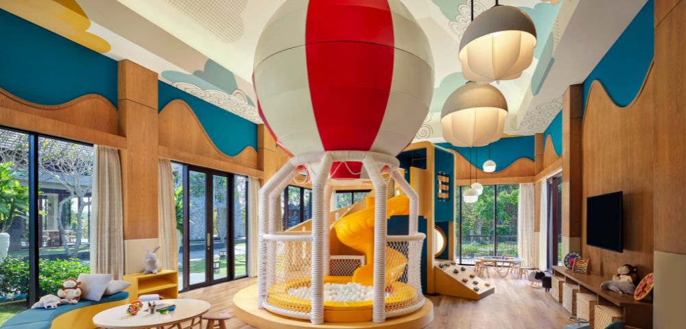 Bright indoor playroom featuring a large balloon-themed slide, colorful decor, wood flooring, round tables, and clouds on the ceiling, creating a playful atmosphere.