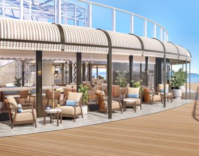 Outdoor cafe with wicker chairs and tables under a striped awning, facing a clear blue sky and ocean. Relaxed ambiance with lush greenery.