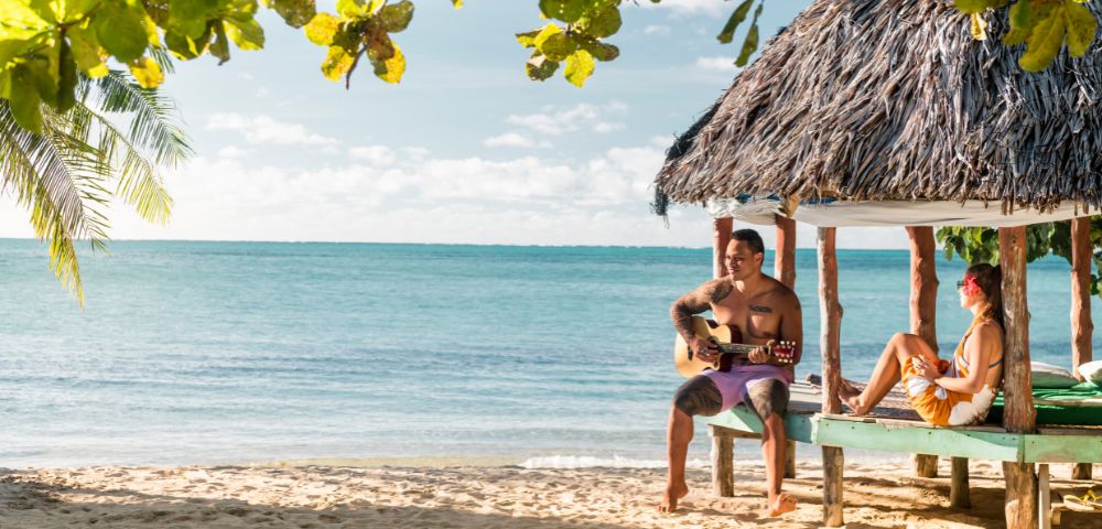 Two people relax on a beach under a thatched hut, enjoying the sun and ocean breeze.