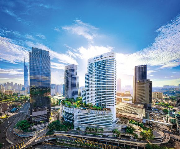 Aerial view of modern cityscape with tall skyscrapers under a bright blue sky. Sunlight breaks through clouds, creating a vibrant and dynamic urban atmosphere.