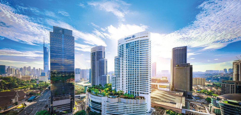 Aerial view of modern cityscape with tall skyscrapers under a bright blue sky. Sunlight breaks through clouds, creating a vibrant and dynamic urban atmosphere.