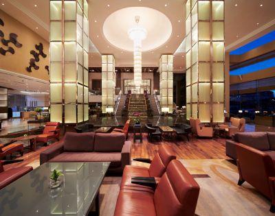 Luxurious hotel lobby with modern decor, featuring a grand staircase, tall illuminated columns, and plush seating. Warm lighting creates an inviting ambiance.