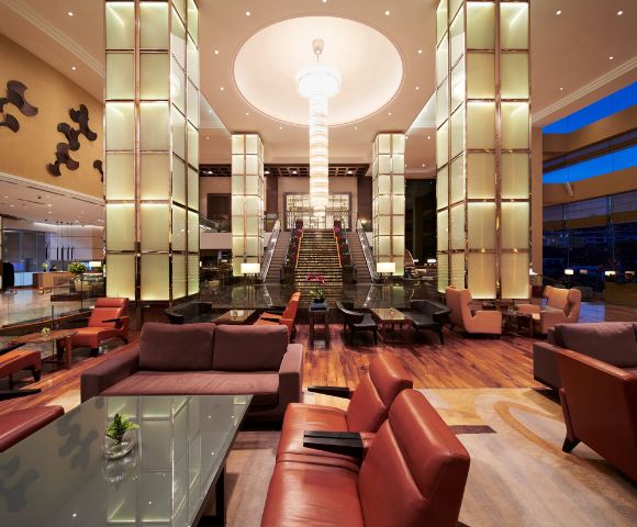 Luxurious hotel lobby with modern decor, featuring a grand staircase, tall illuminated columns, and plush seating. Warm lighting creates an inviting ambiance.