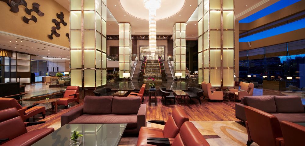 Luxurious hotel lobby with modern decor, featuring a grand staircase, tall illuminated columns, and plush seating. Warm lighting creates an inviting ambiance.