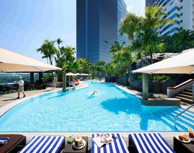 Rooftop pool scene with palm trees, blue water, striped lounge chairs, and sun umbrellas. A person swims, while others relax, conveying a tranquil, resort vibe.