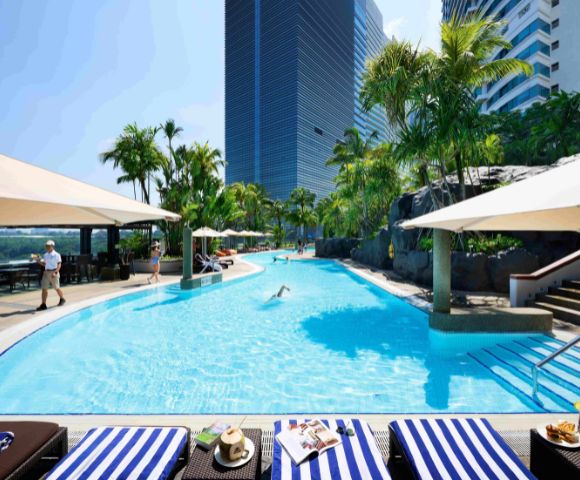 Rooftop pool scene with palm trees, blue water, striped lounge chairs, and sun umbrellas. A person swims, while others relax, conveying a tranquil, resort vibe.