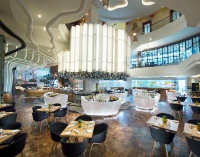 Modern restaurant interior with high ceilings, large white chandelier, curvy architectural features, wooden floors, and elegantly set tables with black chairs.