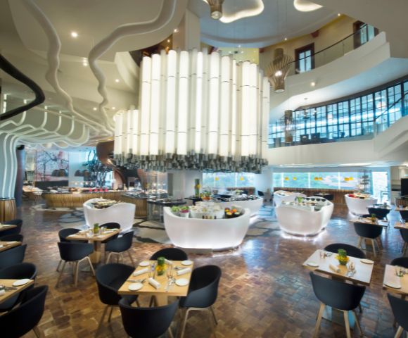 Modern restaurant interior with high ceilings, large white chandelier, curvy architectural features, wooden floors, and elegantly set tables with black chairs.