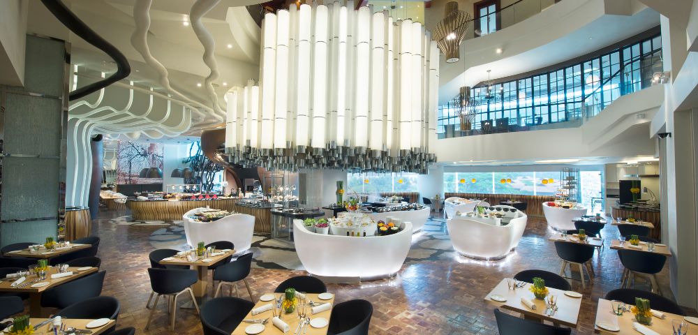 Modern restaurant interior with high ceilings, large white chandelier, curvy architectural features, wooden floors, and elegantly set tables with black chairs.
