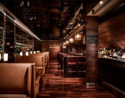 Warm, upscale bar interior with dim lighting, polished wood floors, leather booth seating on the left, and a well-stocked bar on the right. Cozy ambiance.