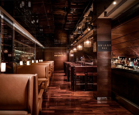 Warm, upscale bar interior with dim lighting, polished wood floors, leather booth seating on the left, and a well-stocked bar on the right. Cozy ambiance.