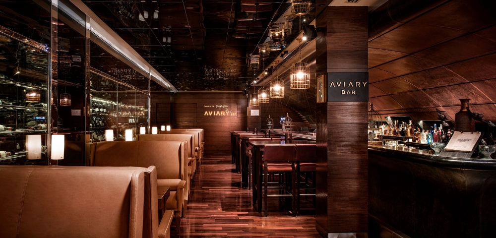 Warm, upscale bar interior with dim lighting, polished wood floors, leather booth seating on the left, and a well-stocked bar on the right. Cozy ambiance.