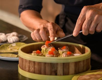 Hands meticulously arrange dim sum topped with roe in a bamboo steamer. In the background, dumplings on a plate suggest a culinary preparation scene.