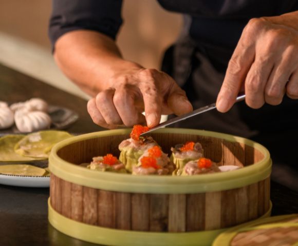 Hands meticulously arrange dim sum topped with roe in a bamboo steamer. In the background, dumplings on a plate suggest a culinary preparation scene.