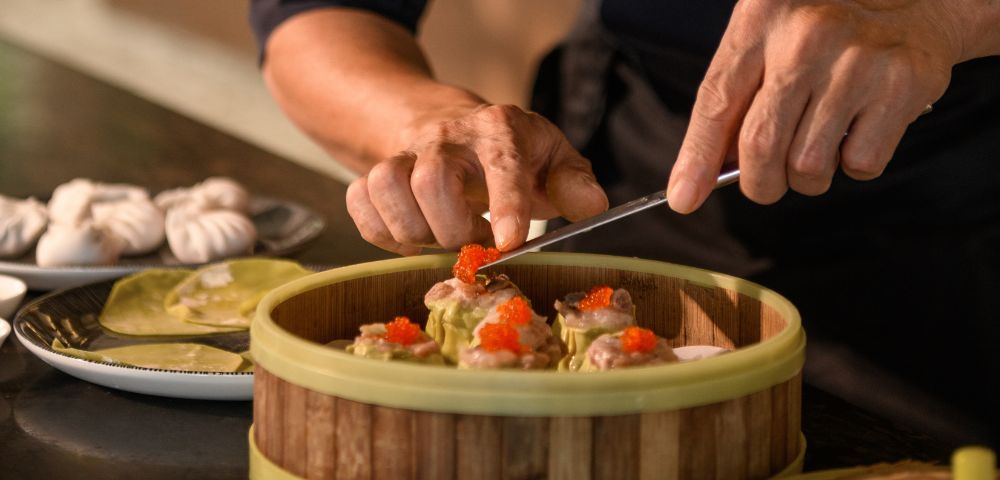 Hands meticulously arrange dim sum topped with roe in a bamboo steamer. In the background, dumplings on a plate suggest a culinary preparation scene.