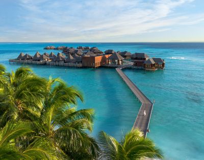 Overwater bungalows on stilts stretch across turquoise sea, connected by a long wooden pier. Palm trees frame the foreground, evoking a tranquil, tropical vibe.